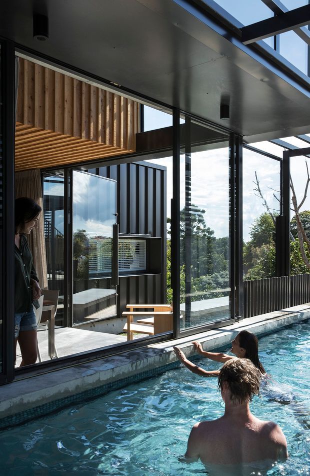 A modern residential pool area viewed from the swimming pool, with swimmers in the foreground and a glazed living space beyond. The architecture features extensive black-framed floor-to-ceiling glazing, a covered outdoor roof structure with exposed dark beams, timber slatted ceiling and wall detailing, and a compact built-in kitchen or bar visible inside. Trees and sky are visible through the glass.