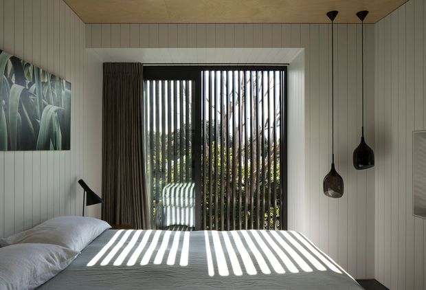 A calm, contemporary bedroom with a neatly made bed positioned in front of full-height glazing. Vertical external or internal screening casts strong striped sunlight across the white bedding. The room features vertically panelled light walls and ceiling, dark floor-length curtains, three black pendant lights, a black bedside lamp, and a botanical wall artwork. Lush planting is visible beyond the screened window.