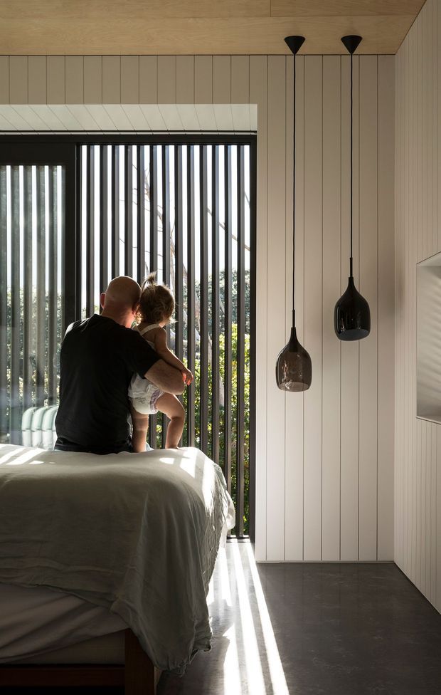 A sunlit contemporary bedroom with a large bed in the foreground, vertical timber wall panelling, a warm timber ceiling, dark-framed floor-to-ceiling glazing screened by vertical black fins, and two sculptural pendant lights. A seated adult holding a child is visible on the bed. The room has a restrained neutral palette and strong linear sunlight across the dark floor.