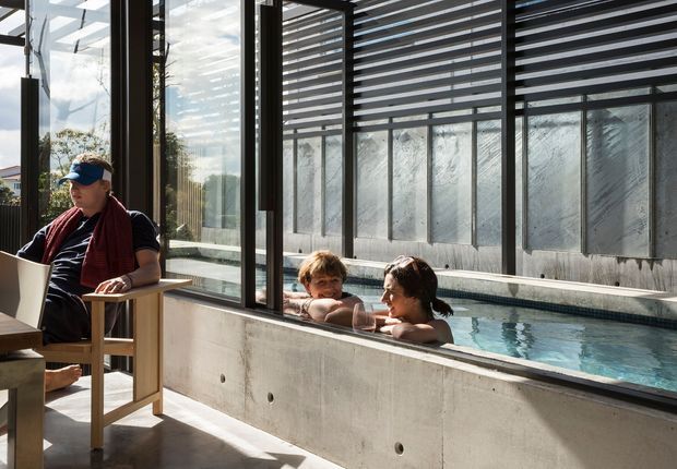 Sunlit contemporary indoor pool area with a narrow lap pool enclosed by full-height glazing and dark horizontal louvres. Exposed concrete walls and floor create an industrial-modern architectural setting. Two swimmers are in the pool, while a seated person works at a table beside the glazing.