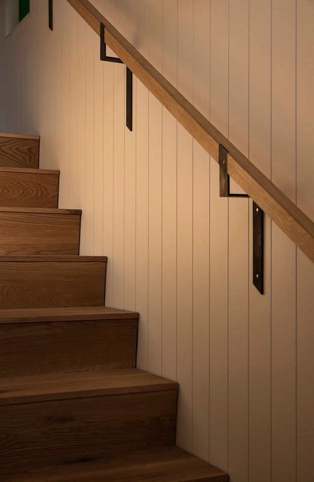 Close-up interior view of a timber staircase with dark wood treads and risers, a warm wood handrail, black metal brackets, and vertical cream wall panelling. The composition highlights the stair detail and contrasting natural materials in a softly lit, refined setting.
