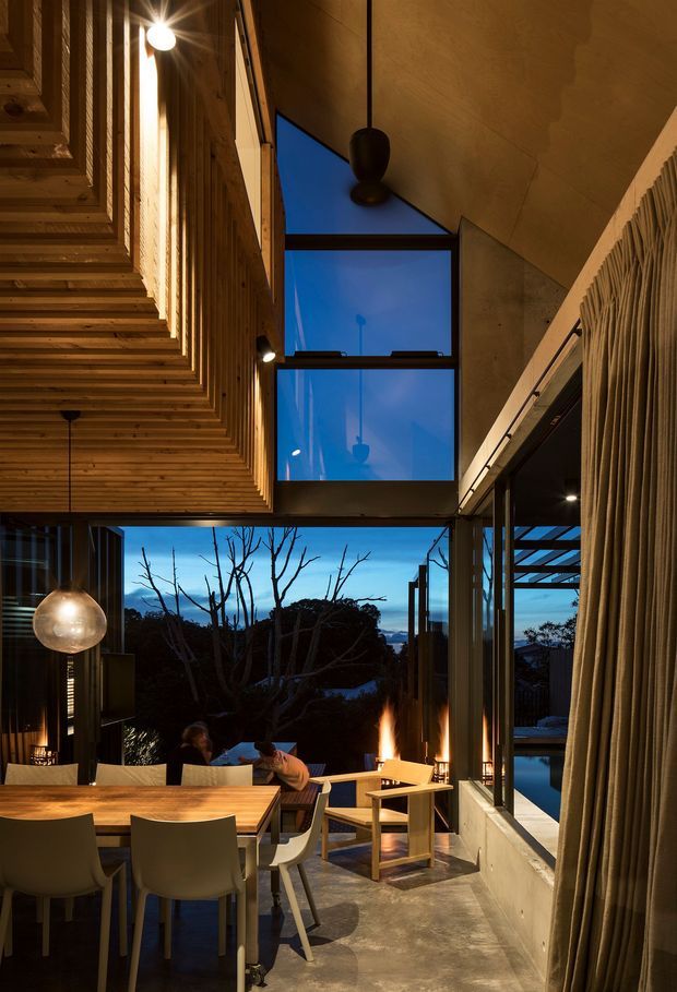 A dramatic double-height dining and living space photographed at dusk. The room features a large timber dining table with contemporary chairs, a concrete floor, extensive floor-to-ceiling glazing, exposed timber ceiling and wall structure, a suspended pendant light, curtains, and a visible indoor fireplace. Large windows open toward trees and a distant landscape, while a sculptural timber-lined ceiling and blue evening light create a warm, intimate atmosphere.