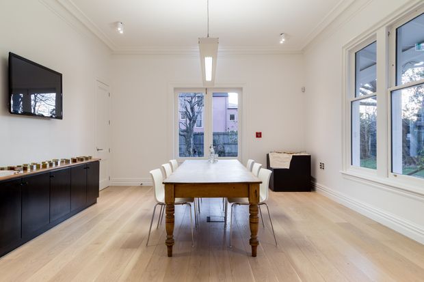 Bright, refined dining room with a traditional architectural shell and contemporary furnishings. The room features a large timber dining table with white upholstered chairs, pale timber flooring, white walls with decorative cornices and trim, a long black built-in sideboard, wall-mounted television, pendant and recessed lighting, and large white-framed windows overlooking greenery.