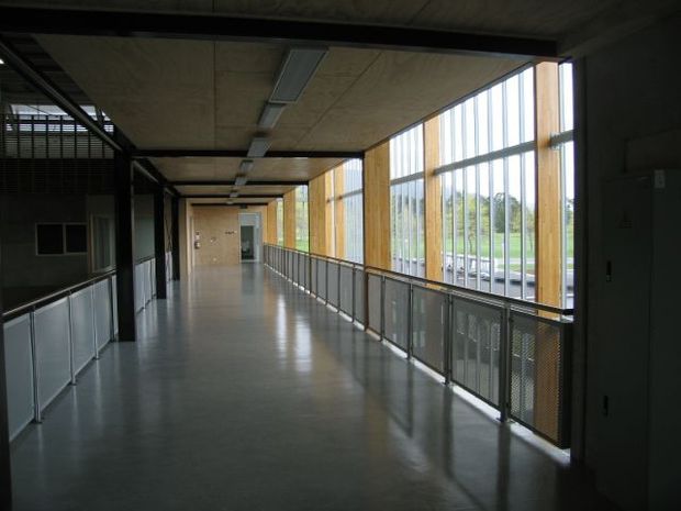 A long, modern institutional corridor or internal walkway with polished grey flooring, exposed concrete ceiling panels, dark structural framing, metal mesh balustrades, and extensive floor-to-ceiling glazing framed in warm timber. The corridor is sparsely furnished and brightly daylit from the windows, with a green outdoor landscape visible beyond.