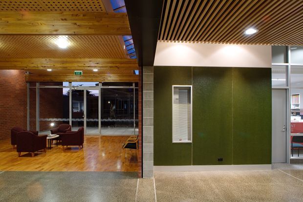 Interior view of a contemporary commercial building lobby or corridor. The space features warm timber parquet flooring, exposed reddish brick walls, a perforated timber ceiling and a slatted timber feature ceiling, with dark green wall panelling, aluminium-framed glazed doors and sidelights, and a small seating area visible in the adjoining lobby. The atmosphere is warm, refined and professional.
