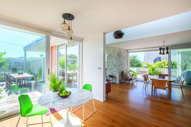 Bright open-plan interior combining a dining area and living room, with extensive glazing opening to a covered outdoor terrace. The space features warm timber flooring, white walls and ceiling, a round marble-topped dining table, vivid green chairs, sculptural pendant lighting, and a textured stone feature wall in the living area. The room has an airy mid-century-influenced contemporary character and strong indoor-outdoor connection.