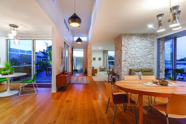 Open-plan residential interior featuring a dining area in the foreground, a living area beyond, and a glazed sitting or breakfast nook to the left. Warm timber flooring, white walls and ceilings, exposed light-coloured stone feature walls, extensive windows, and sculptural pendant lighting create a bright, relaxed contemporary space.