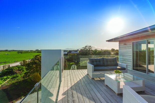A contemporary rooftop or elevated timber deck terrace beside a light weatherboard-clad building, furnished with a grey outdoor sofa and wicker seating. Glass balustrades frame expansive green fields and distant mountain scenery under a bright blue sky.