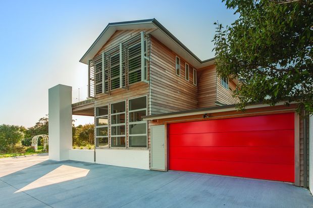Exterior view of a contemporary two-storey home with warm horizontal timber cladding, a prominent red sectional garage door, large glazed entry and upper-level louvred screening. The building has a pitched roof with broad eaves, a concrete driveway, and a small landscaped area visible to the side.