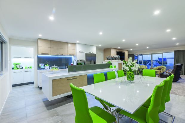 Bright open-plan contemporary kitchen and dining area with a large white island, integrated appliances, light timber cabinetry, glossy white surfaces, and vivid lime-green dining chairs. Recessed ceiling lighting, a dark feature backsplash, and expansive glazing create a clean, modern entertaining space.