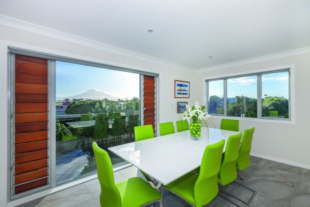 Bright contemporary dining room with a large white rectangular dining table surrounded by vivid lime-green chairs. Floor-to-ceiling glazing and a timber louvred sliding door open to a leafy outdoor view, with a distant mountain visible beyond. The room has pale walls, recessed ceiling lights, grey tiled flooring, and simple framed artwork.