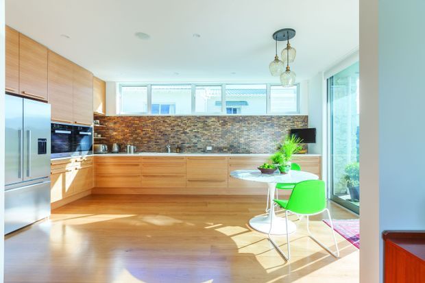Bright contemporary kitchen with extensive warm timber cabinetry, a long stone island or peninsula, stainless-steel appliances, and a multicoloured natural-stone or brick-look splashback. Large windows and glazed doors bring in abundant daylight, with a small dining setting featuring vivid green chairs.