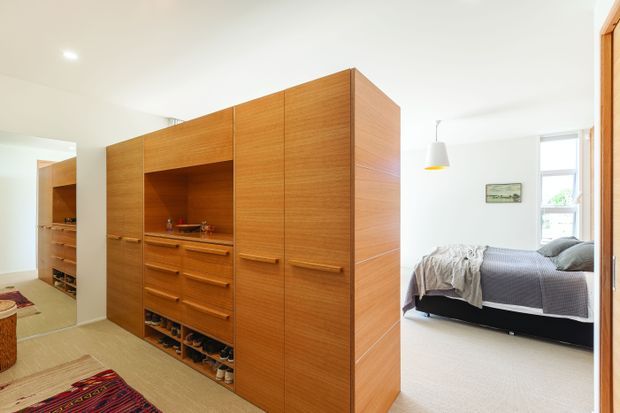 Bright contemporary bedroom area with extensive full-height timber built-in cabinetry and an open central storage niche containing drawers, shelves, and shoes. A partially visible bed with grey bedding sits beyond the cabinetry, while a carpeted floor, white walls, simple pendant light, and daylight create an uncluttered interior.