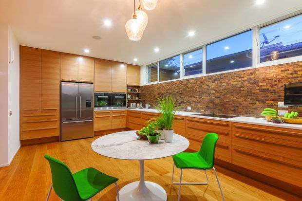 A bright, spacious contemporary kitchen with extensive warm timber cabinetry, integrated appliances, a stainless-steel refrigerator, and a long white countertop. A full-height exposed brick splashback runs beneath clerestory windows, while a round marble-topped dining table with vivid green chairs occupies the foreground. Recessed lighting and pendant lights illuminate the warm timber floor and open-plan kitchen-dining setting.