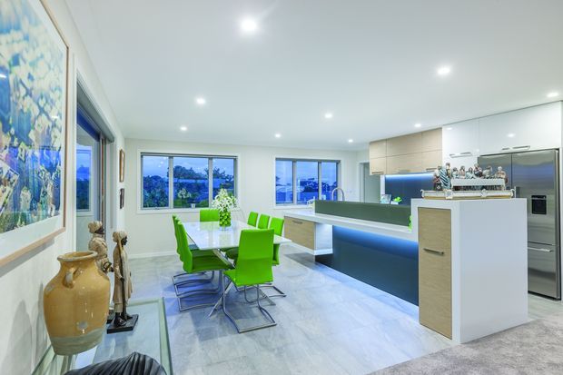 Bright open-plan kitchen and dining area with a contemporary white and light-wood fit-out. The space features a large island with a dark benchtop and blue underlighting, glossy white cabinetry, integrated stainless-steel appliances, pale stone-look flooring, recessed ceiling lights, and a glass dining table with vivid green chairs. Large windows bring in abundant daylight and overlook greenery.