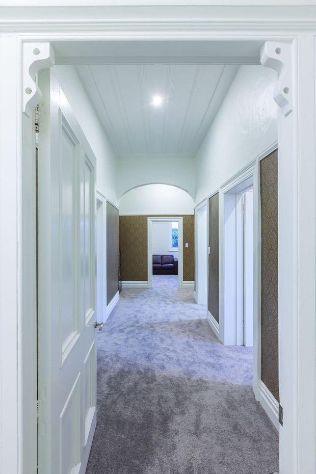 A bright, narrow traditional hallway viewed through an open white paneled door. The corridor has plush grey carpet, white decorative wall and door trim, patterned brown wallpaper panels, a beadboard-style ceiling, and an arched opening leading toward a small sitting area with a dark sofa. Ornate bracket details frame the doorway.