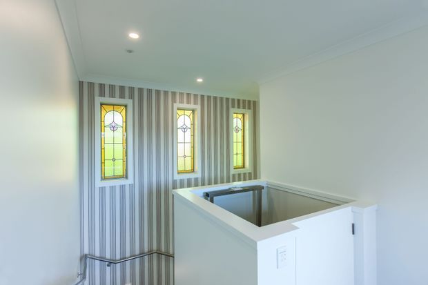 Bright interior stair landing with a white balustrade overlooking a staircase. The far wall features three narrow stained-glass windows with yellow and green panels, set into vertically striped wallpaper. Recessed ceiling lights and simple white trim create a clean, traditional residential feel.