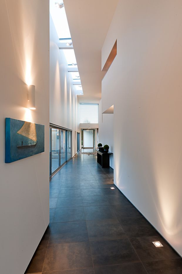 A long, narrow contemporary interior corridor with very high ceilings and a continuous row of skylights. Smooth white walls are punctuated by recessed niches and concealed wall lighting, while large dark grey floor tiles extend through the space. Full-height glazed doors or windows line one side, and a small dark console with spherical decor sits near the far end. The corridor has a bright, refined gallery-like atmosphere with strong linear perspective.