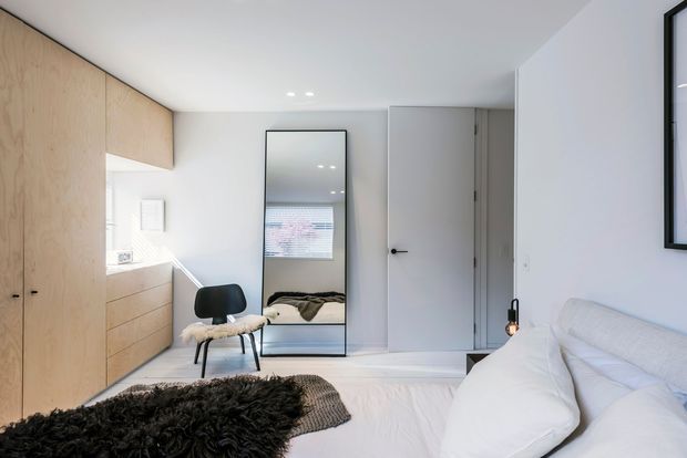 A compact, bright bedroom with a minimalist fit-out. The room contains a low bed with white bedding, pale timber built-in cabinetry along the left wall, a small black chair, a full-height leaning mirror, and a simple white door. White walls and ceiling, light timber joinery, and restrained furnishings create a clean, calm atmosphere.