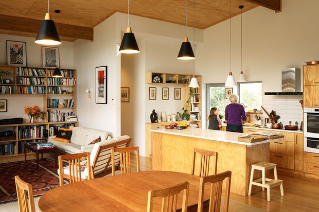 Warm open-plan kitchen, dining, and living area with extensive timber finishes. The space features a large timber kitchen island, timber cabinetry, a dining table with wooden chairs, open bookshelves, a patterned rug, a white sofa, exposed timber ceiling boards and beams, and multiple black pendant lights. The kitchen and living areas are visually connected, with daylight entering through the rear windows.