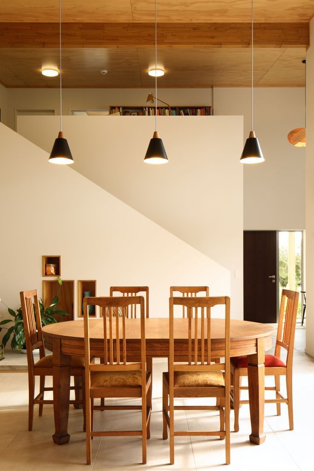 A warm, light-filled dining room with a large oval timber dining table and matching high-back timber chairs. The room features cream painted walls, a dramatic diagonal stair or partition wall, a timber-lined ceiling with exposed timber beam detailing, three black pendant lights, recessed ceiling lights, built-in shelving with books, and a dark timber entry door. A small plant and framed objects add restrained decoration.