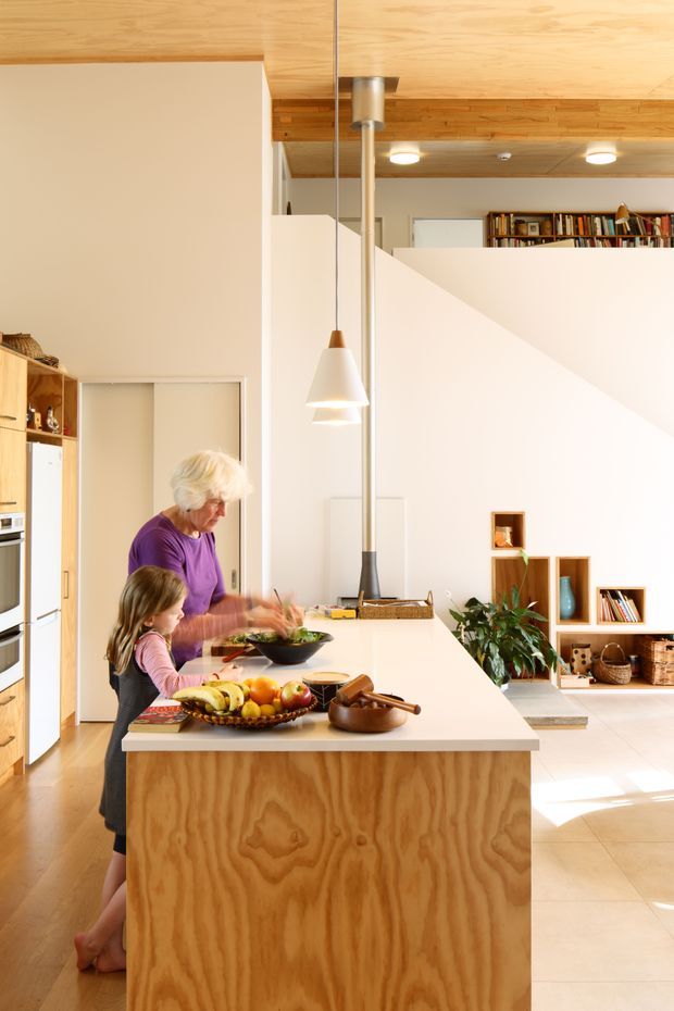 Bright open-plan kitchen centred on a large white island with a plywood-clad base and integrated cooking area. The space features extensive warm timber ceiling lining and cabinetry, white walls, a sculptural pendant light, open shelving, and a partially visible living or reading area beyond the kitchen. A stepped partition and high ceiling create an airy, contemporary family interior.