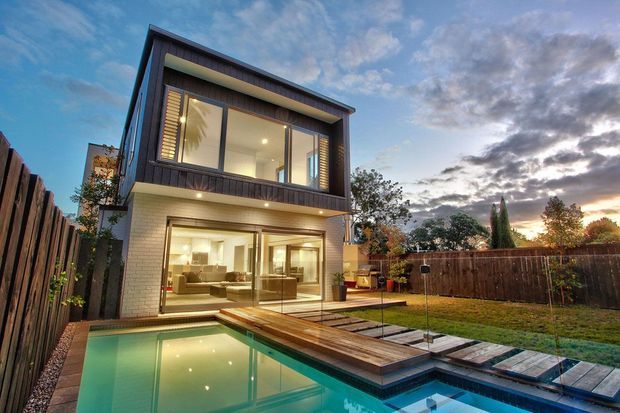 Contemporary two-storey residential rear exterior at dusk, featuring a compact rectangular form with dark vertical upper cladding, light brickwork at ground level, expansive glazing, and a timber-framed balcony or upper terrace. A swimming pool with timber decking occupies the foreground, with a fenced lawn and landscaped suburban garden beyond.