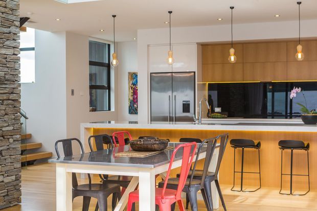 Open-plan contemporary kitchen and dining area with a large island breakfast bar, timber cabinetry, black splashback, stainless-steel refrigerator, pendant lights, and a dining table with mixed metal chairs. A stacked-stone feature wall and stair edge are visible at the left.