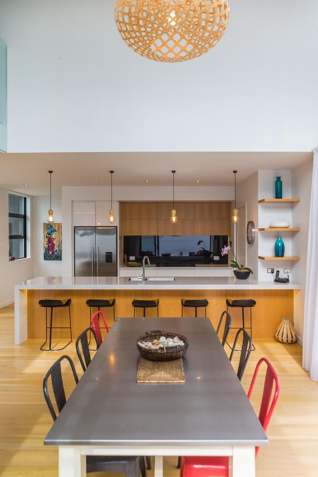 Bright open-plan kitchen and dining area with a central island and breakfast bar, timber cabinetry, stainless-steel appliances, dark benchtops, pendant lighting, and a large dining table. The space has light timber flooring, white walls, open shelving with decorative objects, and colourful accents.
