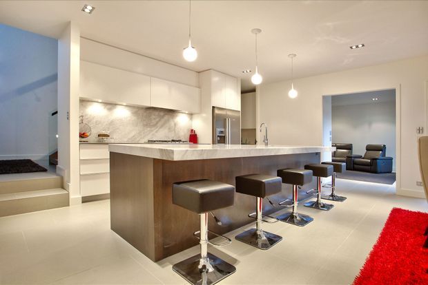 Bright open-plan contemporary kitchen dominated by a large stone island with four bar stools. The kitchen has glossy white handleless cabinetry, a marble-look splashback, integrated stainless-steel appliances, pale tiled flooring, pendant lighting, and a lounge area visible beyond the opening.