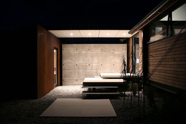 A dark, sophisticated modern courtyard or outdoor lounge viewed at night. The space features a gravel ground plane with large rectangular stepping slabs, a long concrete feature wall with exposed formwork marks, a minimalist floating bench or table, timber-clad walls and extensive dark-framed glazing. Recessed ceiling lights create a warm, dramatic atmosphere, while sparse planting softens the architectural composition.