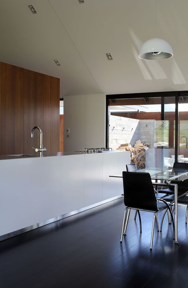 A contemporary open-plan kitchen and dining area with a long white island, integrated cooktop and curved stainless-steel tap, dark timber wall cabinetry, and a black dining table with matching modern chairs. Dark flooring contrasts with the white cabinetry and pale ceiling. Full-height black-framed glazing opens the room to a covered outdoor terrace, with a suspended white pendant light and recessed ceiling downlights.