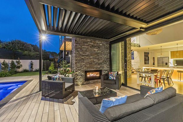A contemporary covered outdoor entertaining area adjoining an open-plan kitchen and dining space. The patio features a timber-look deck, dark slatted pergola roof, stacked-stone feature wall with a built-in fireplace, outdoor lounge furniture, and views toward a landscaped yard and swimming pool. Warm interior lighting contrasts with the blue evening sky.