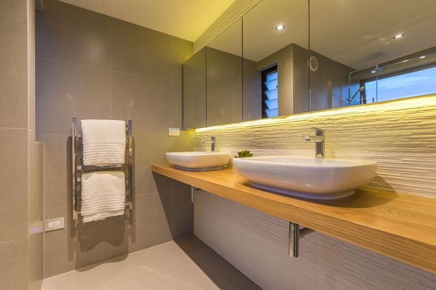 Contemporary bathroom with a double vessel vanity, warm timber benchtop, textured horizontal feature splashback, large mirrored cabinetry, recessed ceiling lights, and a heated towel rail. The neutral grey and warm wood palette creates a refined, softly lit interior.