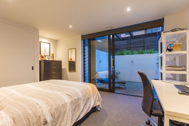 A contemporary bedroom with a neatly made bed, soft carpet flooring, built-in or freestanding dark drawer storage, a white desk and open shelving, and a large glazed sliding door opening toward a small private courtyard. The room is softly lit with recessed ceiling lights and has a calm, neutral palette.