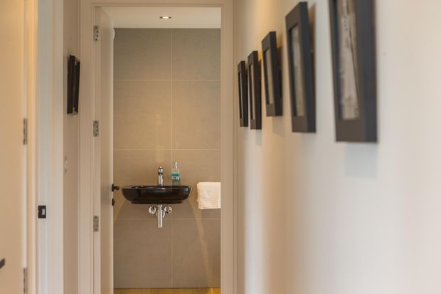 A narrow interior hallway leads directly to a compact bathroom or powder room. The bathroom features large beige wall tiles, a dark wall-mounted basin with exposed plumbing, a chrome tap, a small recessed ceiling light, and a white towel. The hallway walls are painted off-white and decorated with a series of vertically arranged dark-framed artworks.