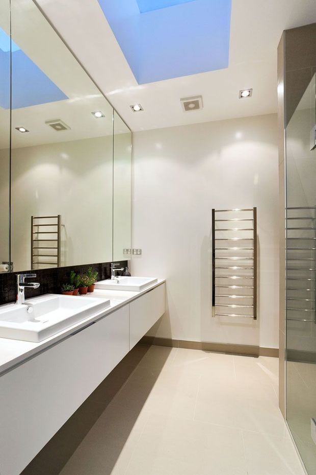 Bright contemporary bathroom with a long floating white vanity, two rectangular basins, a large wall-to-wall mirror, dark tiled splashback, chrome heated towel rail, pale floor tiles, recessed ceiling lights, and a rectangular skylight. A glazed shower enclosure is visible at the right.