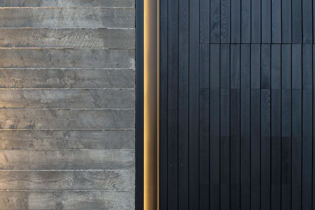 Close-up detail of a contemporary exterior facade combining weathered horizontal grey timber boards on the left with dark vertical timber cladding on the right, separated by a narrow warm metallic or illuminated trim. The composition emphasizes contrasting cladding directions, textures, and dark neutral tones.