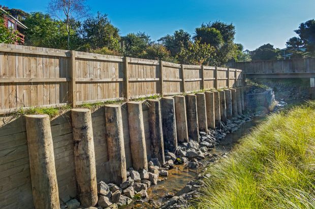 Outdoor view of a timber retaining fence and drainage channel, reinforced with a row of large cylindrical timber posts and rock riprap. Dense vegetation, ornamental grasses, and a small trickle of water run along the base, with a residential property partly visible beyond the fence.