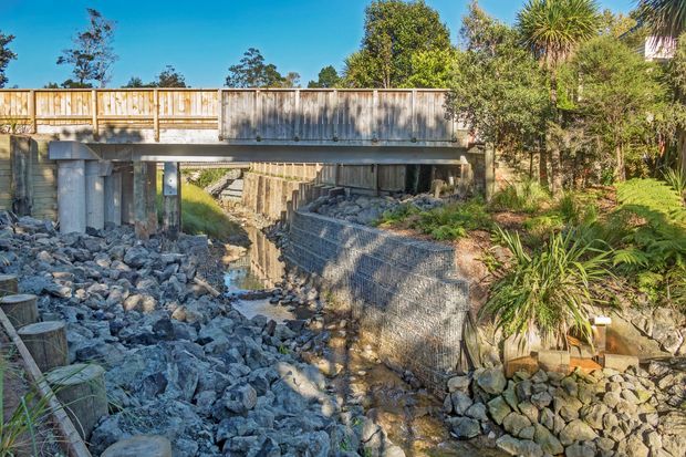 Exterior view of a narrow pedestrian bridge crossing a landscaped rocky stream channel. The bridge has a simple timber-clad railing and grey structural supports, with gabion-style stone retaining walls, boulders, native planting and a small flowing creek beneath. Bright clear daylight and mature vegetation frame the scene.