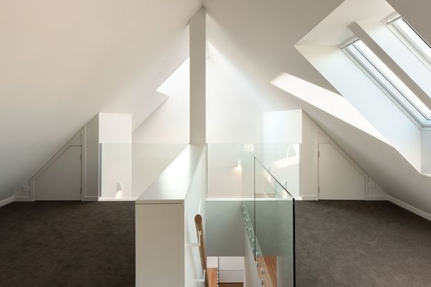 A bright attic-level landing or upper hallway with steeply sloped white ceilings, multiple skylights, and a central stairwell enclosed by clear glass balustrades. White built-in cupboards and concealed storage sit beneath the eaves, while dark grey carpet covers the floor. The space is minimalist and airy, with a glimpse of the stairway and lower level below.