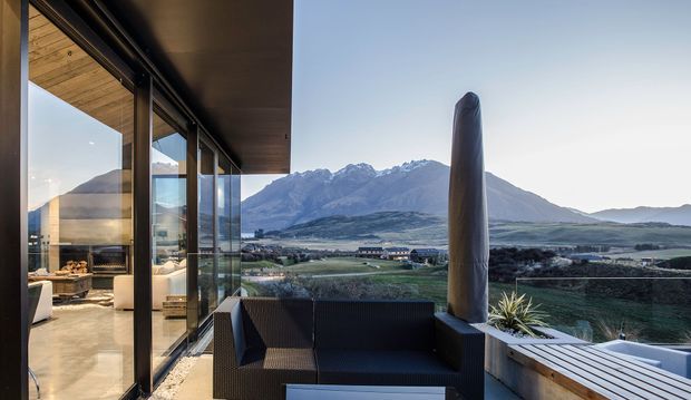 A contemporary covered outdoor terrace or balcony overlooking a dramatic mountain and rural valley landscape. Full-height glazed sliding doors connect the terrace to a warmly finished interior, while dark outdoor seating, timber decking, a raised planter and a sculptural vertical feature create a refined indoor-outdoor setting.