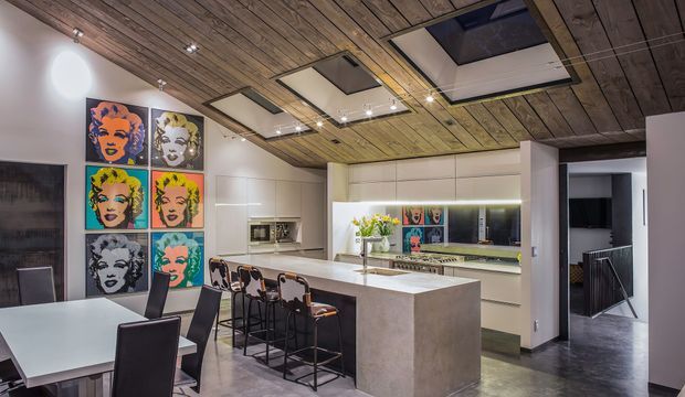 Open-plan contemporary kitchen and dining area with a large pale stone island and integrated breakfast bar, white flat-panel cabinetry, mirrored splashback, stainless appliances, and dark bar stools. The space features a striking timber-planked ceiling with multiple recessed skylight-like ceiling panels and track lighting. A colorful pop-art portrait gallery adds a bold accent beside the dining area, while a hallway and stair zone are visible beyond the kitchen.