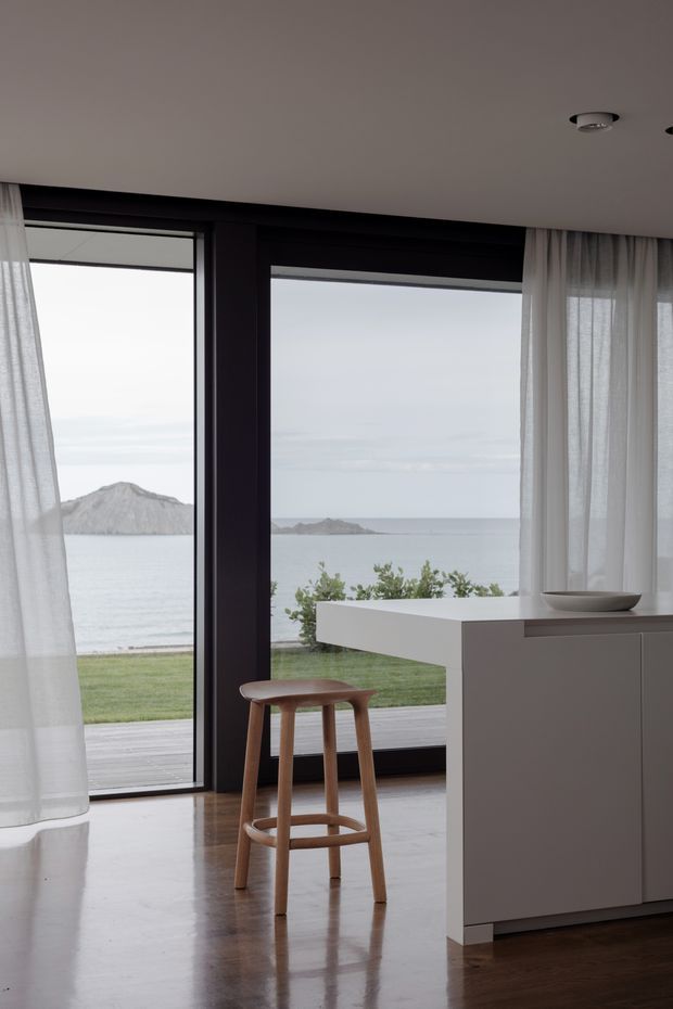A minimalist kitchen interior with a white island and a wooden bar stool, framed by full-height black window and door glazing. Sheer white curtains soften the room, while the polished timber floor reflects the neutral cabinetry. Through the glazing, a calm ocean, rocky offshore islands, lawn and a timber deck are visible.