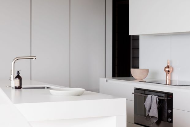 A bright, minimalist contemporary kitchen featuring a white island with integrated sink and slim brushed-metal tap, white handleless cabinetry, a built-in oven, and a dark vertical pantry or shelving recess. Subtle blush-toned accessories and a grey dish towel provide restrained contrast.