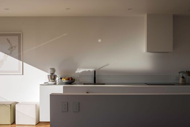 A minimalist contemporary kitchen viewed across a large island or peninsula. The space features smooth white and pale grey surfaces, integrated cabinetry, a dark undermount sink tap, a sleek cooktop with concealed rangehood, and restrained countertop accessories. Warm timber flooring and strong diagonal sunlight create contrast against the clean, neutral joinery.