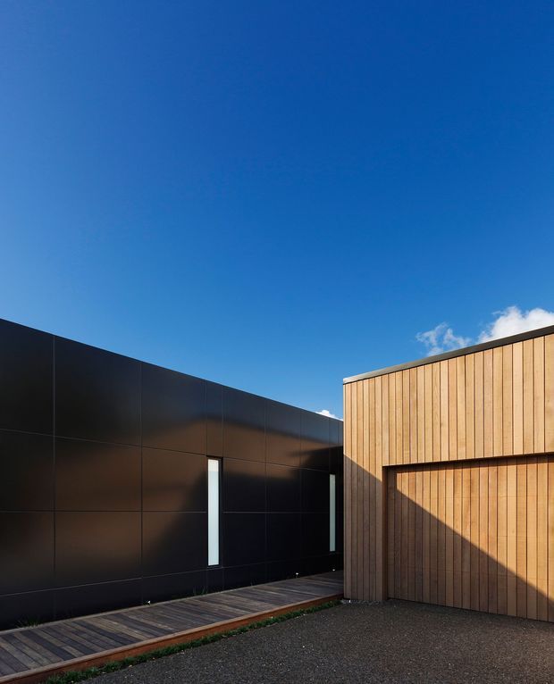 Exterior architectural view of a contemporary building composed of a dark, smooth-panelled wing and a contrasting vertical timber-clad volume. A narrow timber walkway runs alongside the building, with a gravel or paved foreground and a clear blue sky.