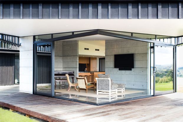 A contemporary indoor-outdoor living space with expansive frameless-style glazed walls opening onto a broad timber deck. The room features exposed concrete walls, dark metal window and door frames, a wall-mounted television, a dining area, and pale lounge furniture. A green landscape is visible beyond the glazing.
