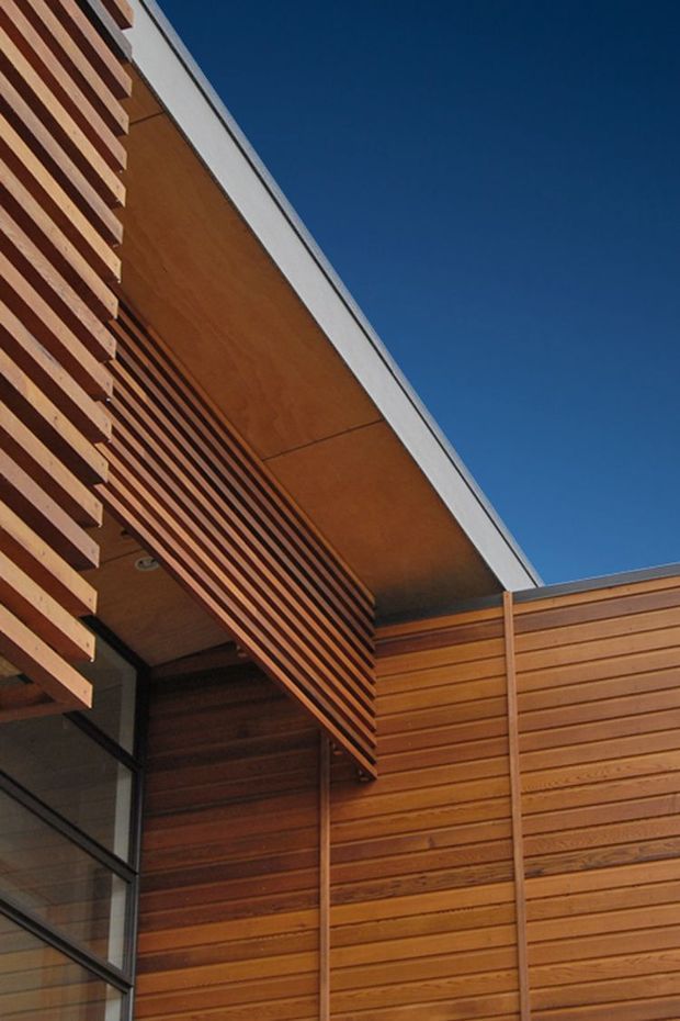 Close-up upward view of a contemporary building exterior featuring warm timber cladding, a projecting roof canopy with timber soffit, horizontal timber slats, and dark-framed glazing against a clear blue sky.