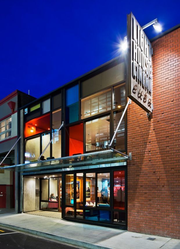 Night-time street-level view of a contemporary multi-storey commercial building with a red brick facade, extensive black-framed glazing, a projecting glass canopy, and brightly illuminated retail or hospitality interiors. A large vertical sign is mounted prominently on the corner facade.